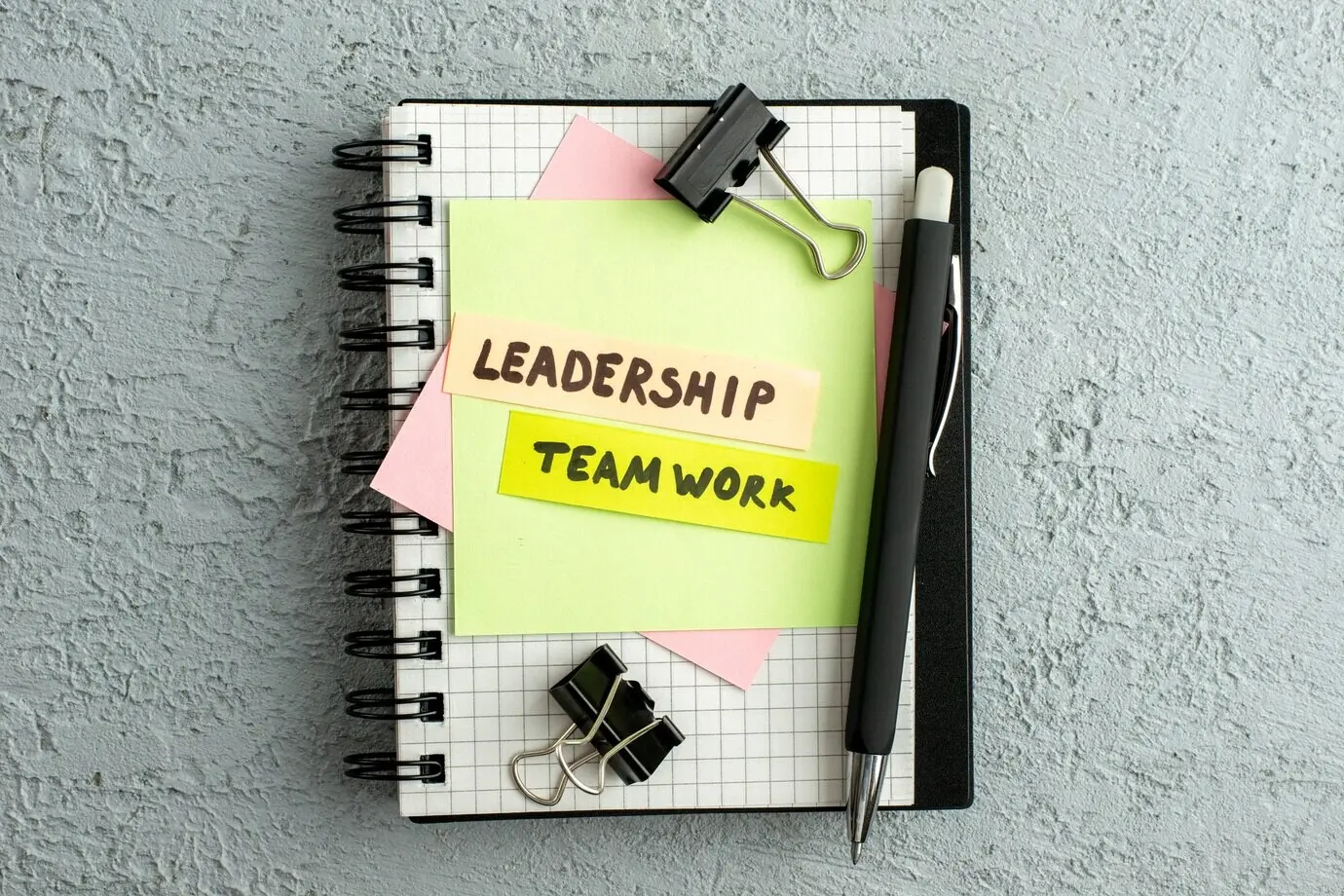 Draufsicht: Führung und Teamarbeit auf farbigen Umschlägen, die auf einem Spiralnotizbuch und einem Buch liegen, vor grauem Sandhintergrund.